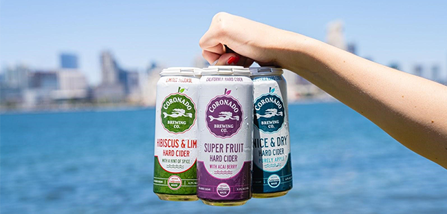 Hand holding three Coronado Brewing hard ciders—Hibiscus & Lime, Super Fruit, and Nice & Dry—with a city skyline and bay in the background, showcasing gluten free cider options in the cider vs beer debate.