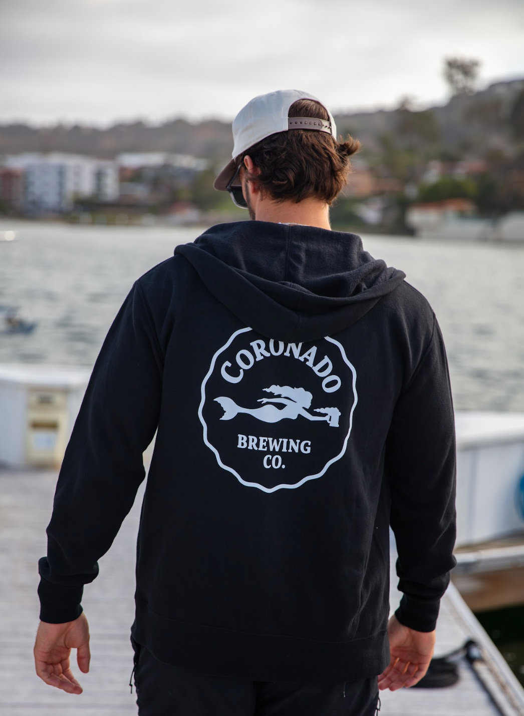 Person wearing a black hoodie with 'Coronado Brewing Co.' logo, standing by a waterfront.