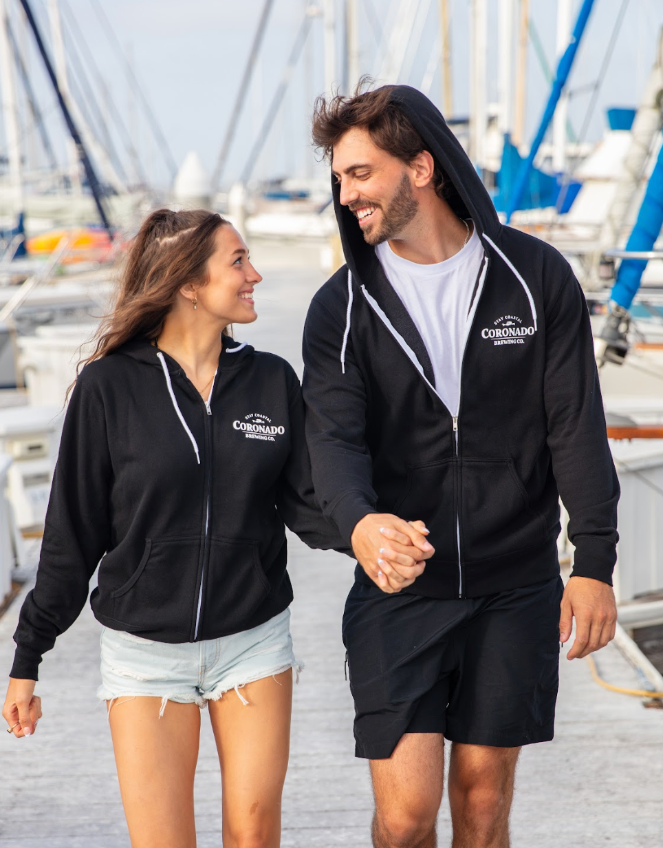 A couple  wearing black Coronado Brewing Co. zip-up hoodies with boats in the background