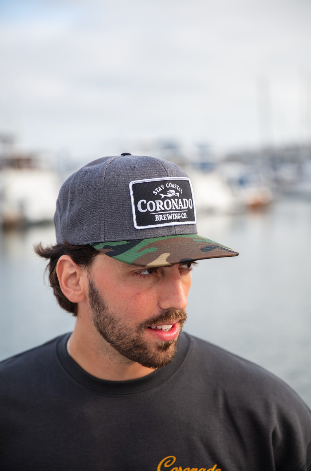 Man wearing a gray and green cap with 'Coronado Brewing Co' logo, standing by water.