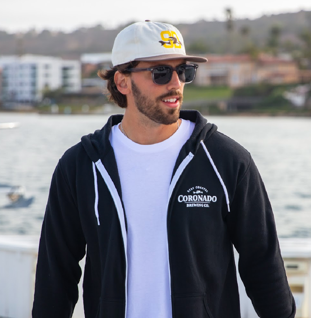 Man wearing a black hoodie with 'Coronado Brewing Co' logo, white t-shirt, and cap outdoors by water.