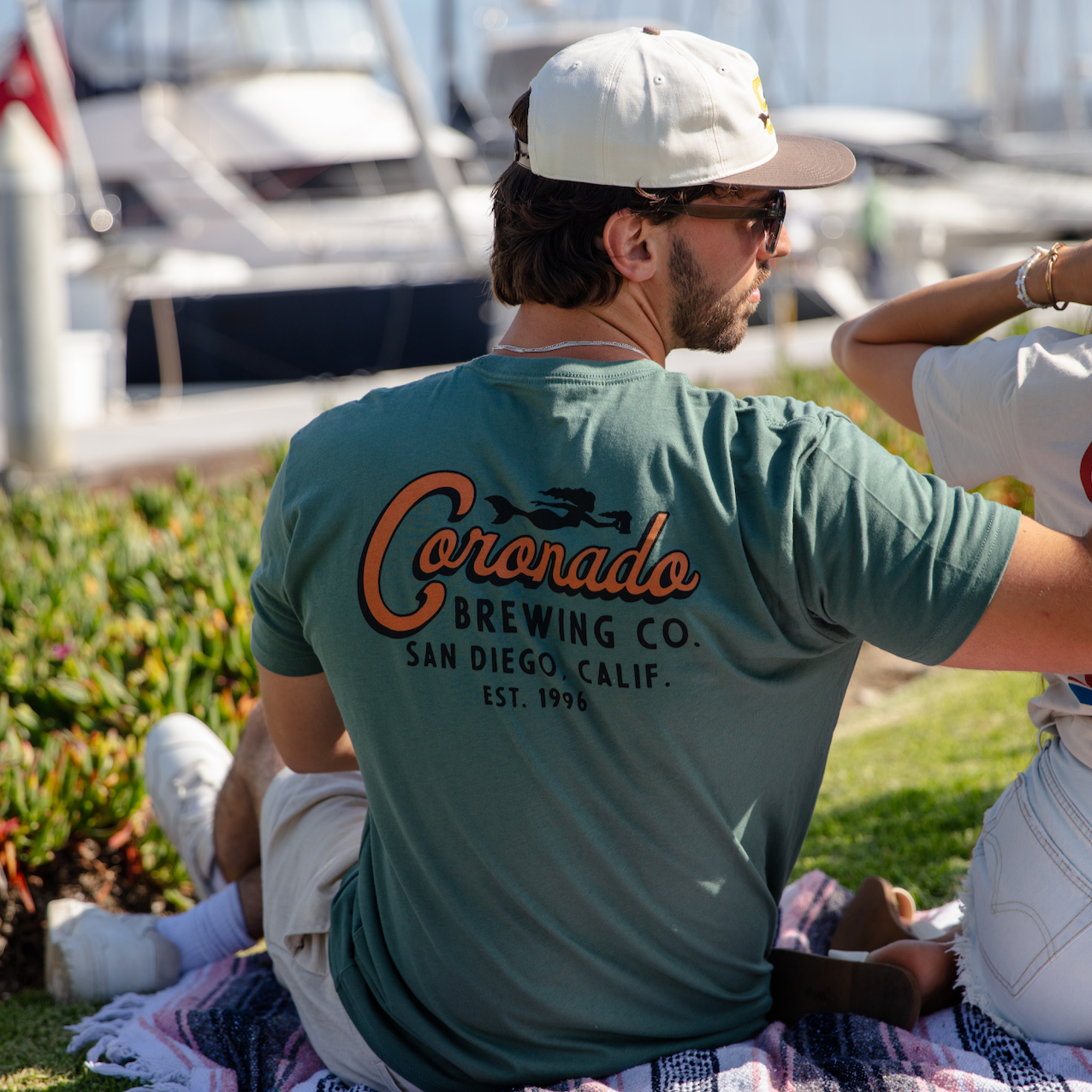 Man sitting on a blanket with a 'Coronado Brewing Co.' t-shirt, surrounded by boats.
