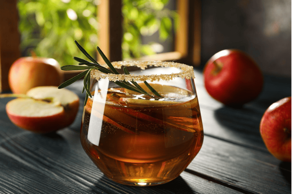 A glass of cider on a dark wooden table surrounded by apples