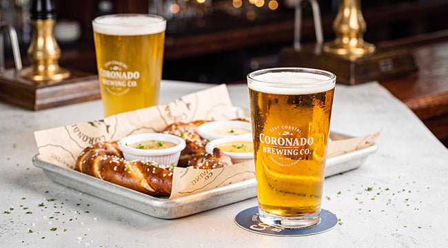 Two Coronado Brewing pints with creamy white foam heads beside a tray of soft pretzels and dipping sauces on a bar