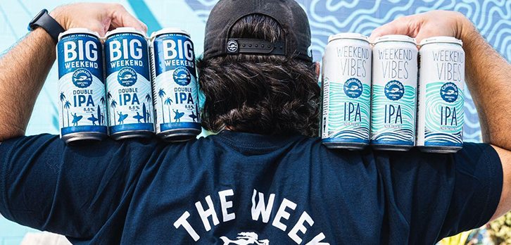 Person holding Big Weekend and Weekend Vibes beer cans in front of “Stay Coastal” mural at Coronado Brewing Co.