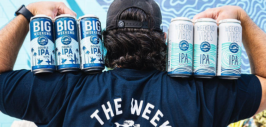 Person holding Big Weekend and Weekend Vibes beer cans in front of “Stay Coastal” mural at Coronado Brewing Co.