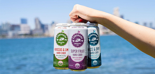 Hand holding three Coronado Brewing hard ciders—Hibiscus & Lime, Super Fruit, and Nice & Dry—with a city skyline and bay in the background, showcasing gluten free cider options in the cider vs beer debate.