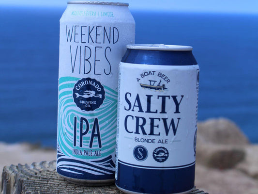 Two cans of Coronado Brewing Co. Weekend Vibes IPA and Salty Crew Blonde Ale clinking together on a wooden post with a beach view, capturing the essence of a San Diego brewery tour experience.