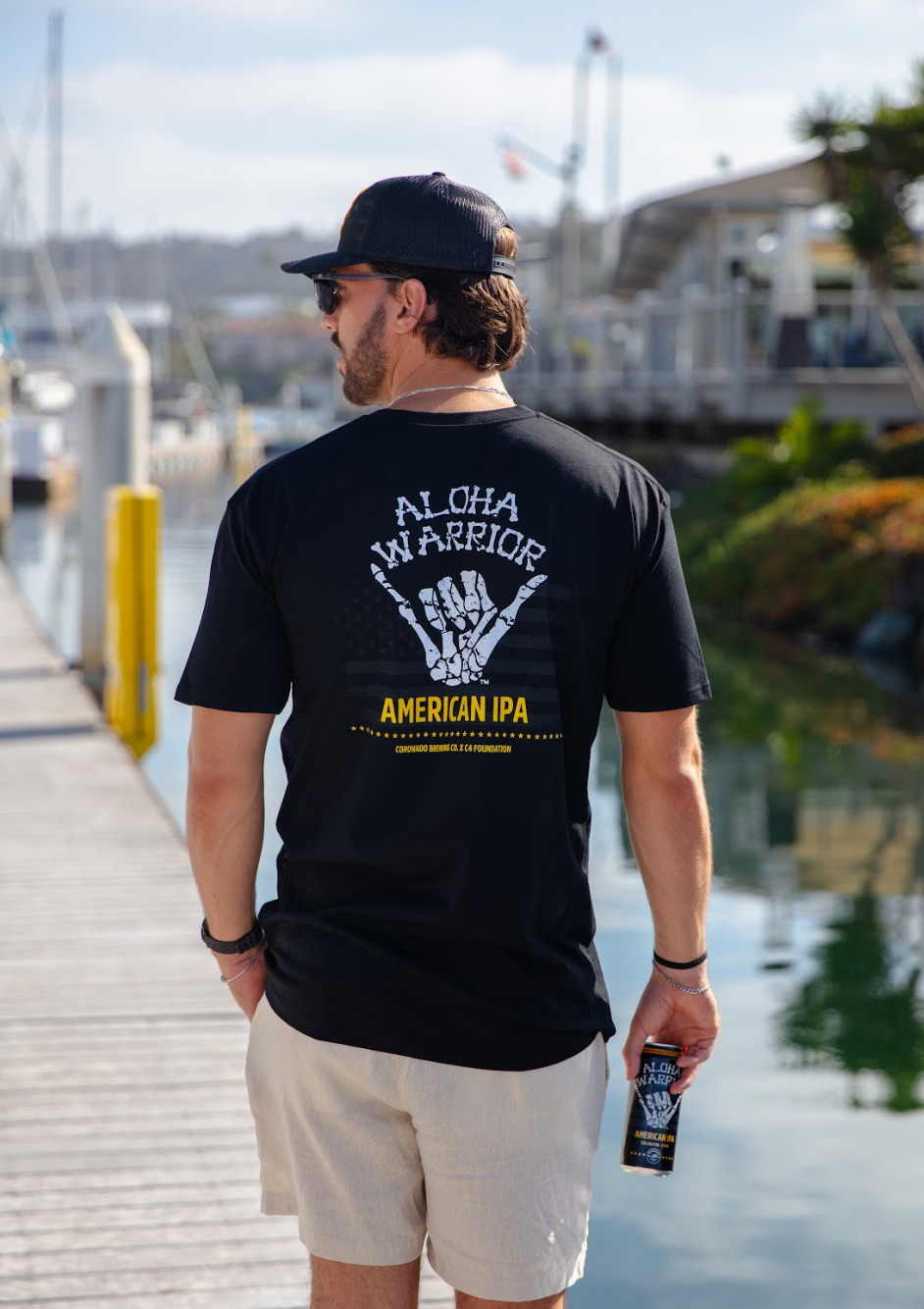 Man wearing a black t-shirt with 'Aloha Warrior American IPA' design, standing on a dock.