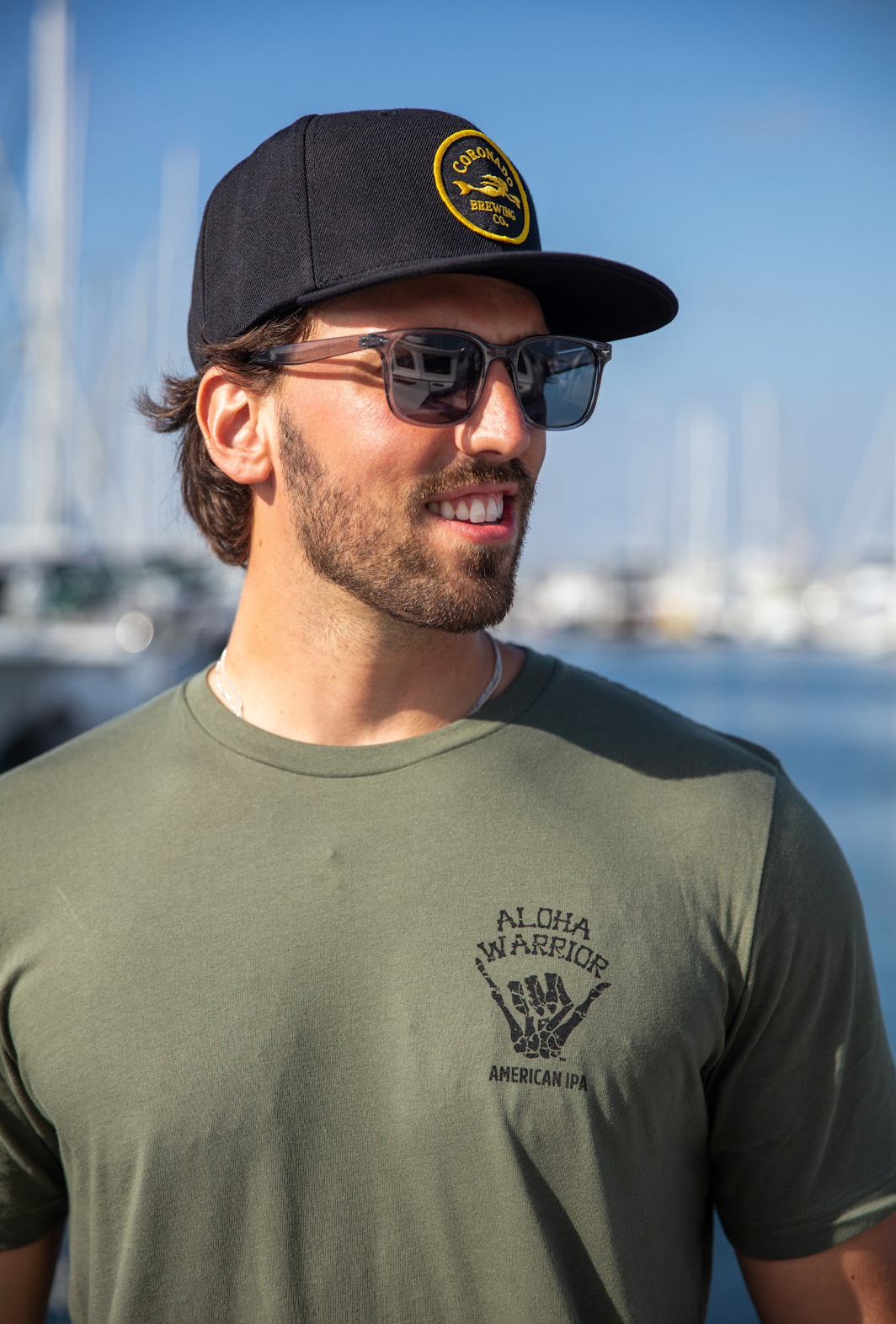 A man wearing a black Coronado Brewing Co. cap and sunglasses with a green Aloha Warrior t-shirt featuring , standing outdoors with boats in the background.
