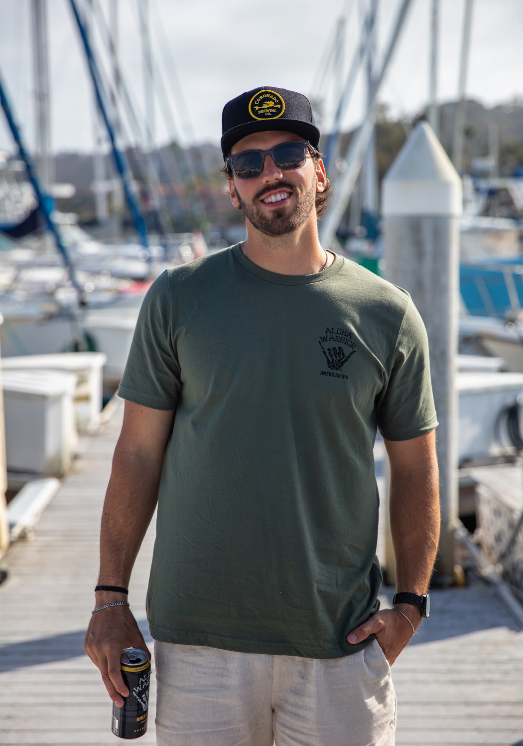 Man wearing a green Aloha Warrior t-shirt, sunglasses, and a Coronado Brewing Co. hat, standing on a dock with boats in the background.