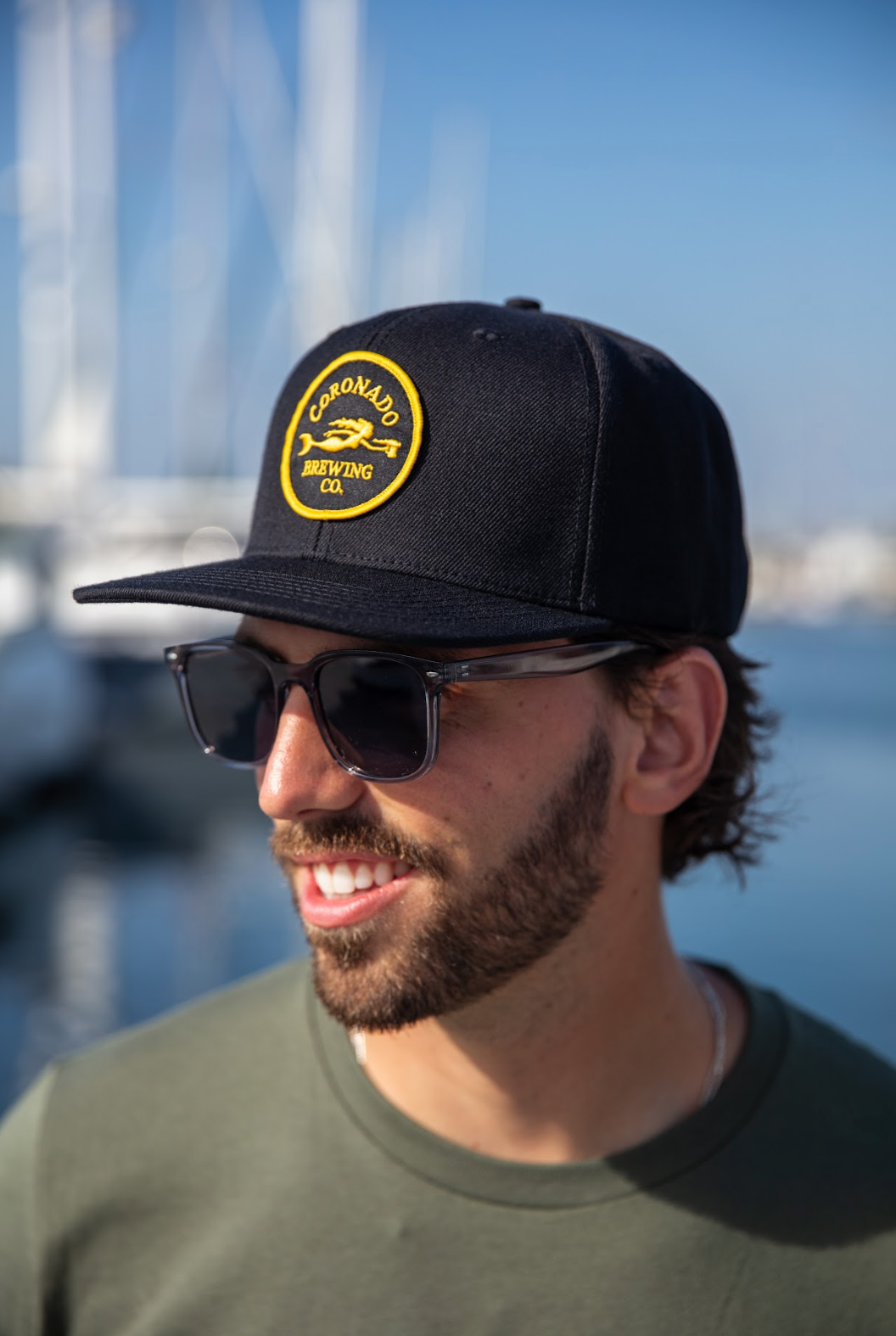 Man wearing a black Coronado Brewing  Co. hat with a yellow logo and sunglasses, standing by water.