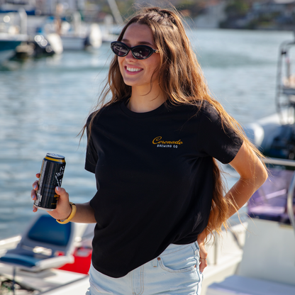 Woman on a boat holding a can, wearing a black t-shirt and denim shorts, with water and boats in the background.