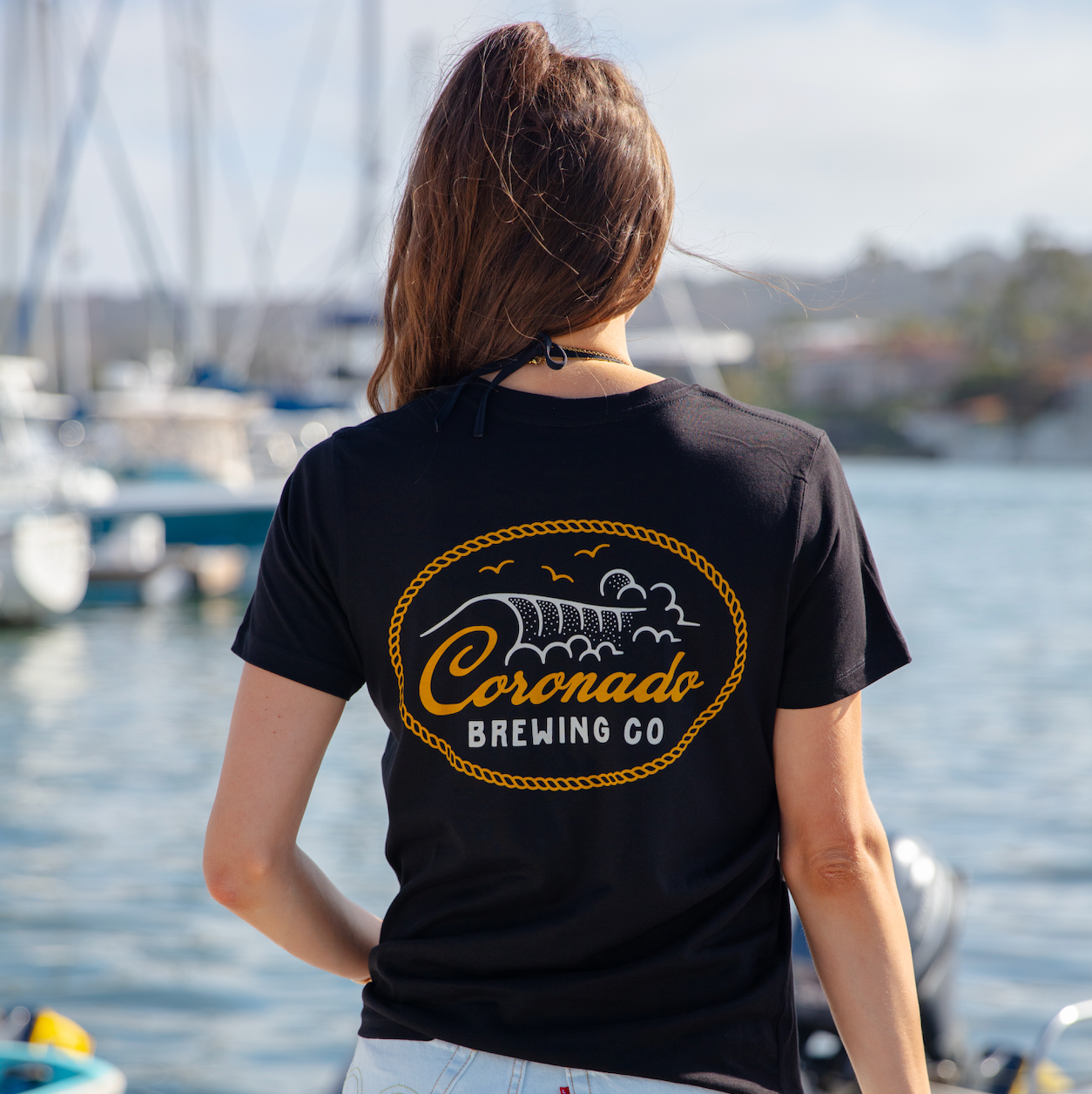 Person wearing a black t-shirt with 'Coronado Brewing Co' logo by a waterfront.