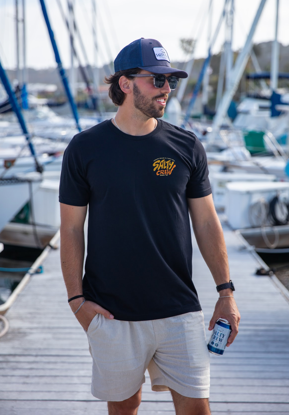 Man wearing a black t-shirt with a logo, white shorts, and a blue cap, standing on a dock with boats in the background.