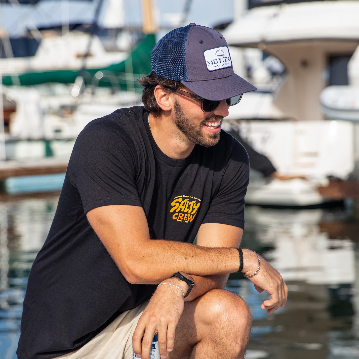 Man wearing a Salty Crew cap and t-shirt sitting by a marina.