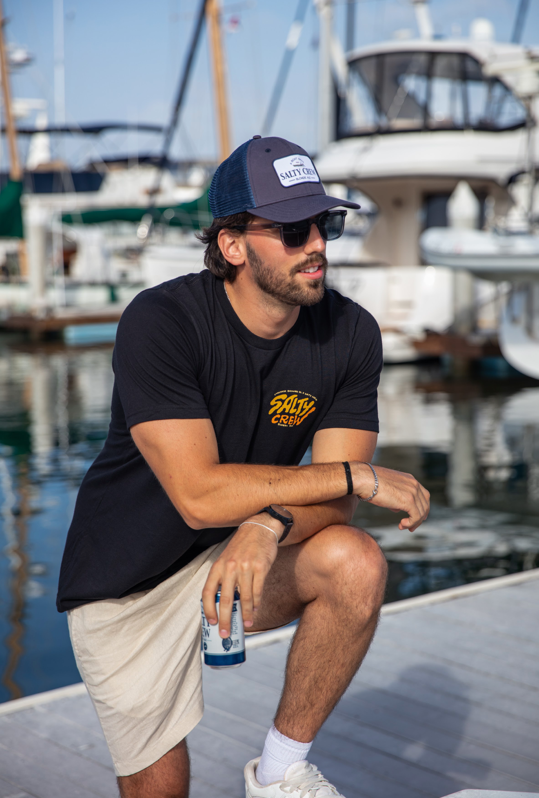 Man sitting on a dock by boats wearing a black t-shirt and Salty Crew Boat Beer hat.
