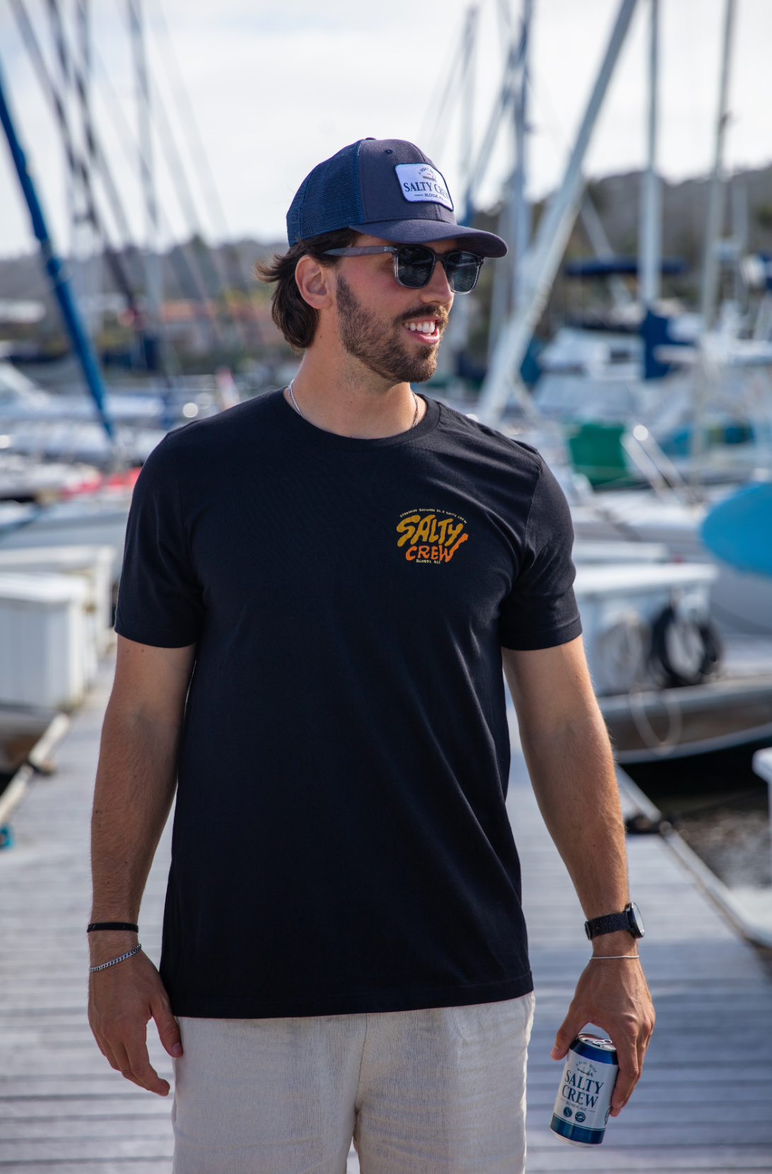 Man wearing a black t-shirt with a logo, sunglasses, and a cap, standing on a dock with boats in the background.