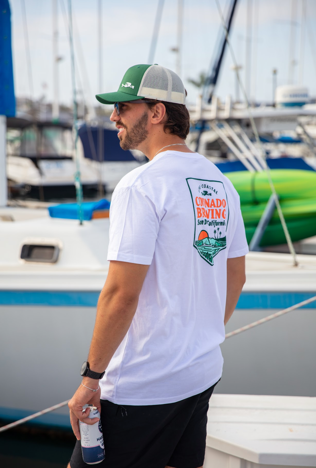 Man wearing a white t-shirt with a  Coronado Brewing logo, standing on a dock with boats in the background