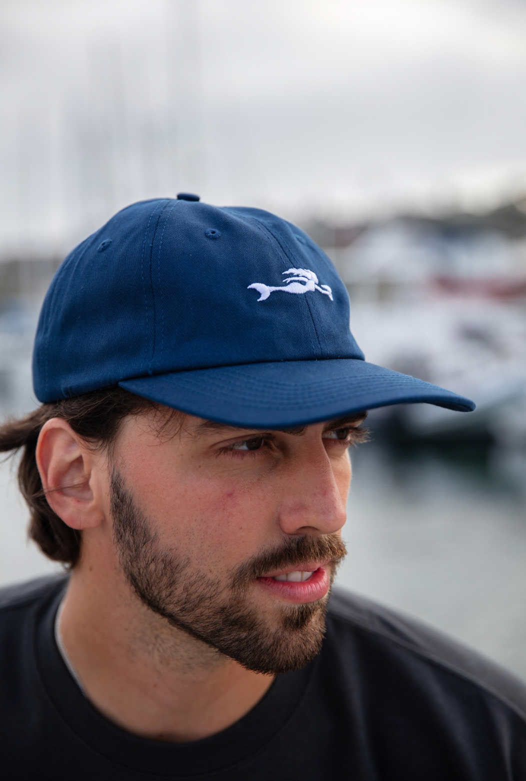 Man wearing a blue Coronado Brewing Co. cap with a mermaid logo, blurred background