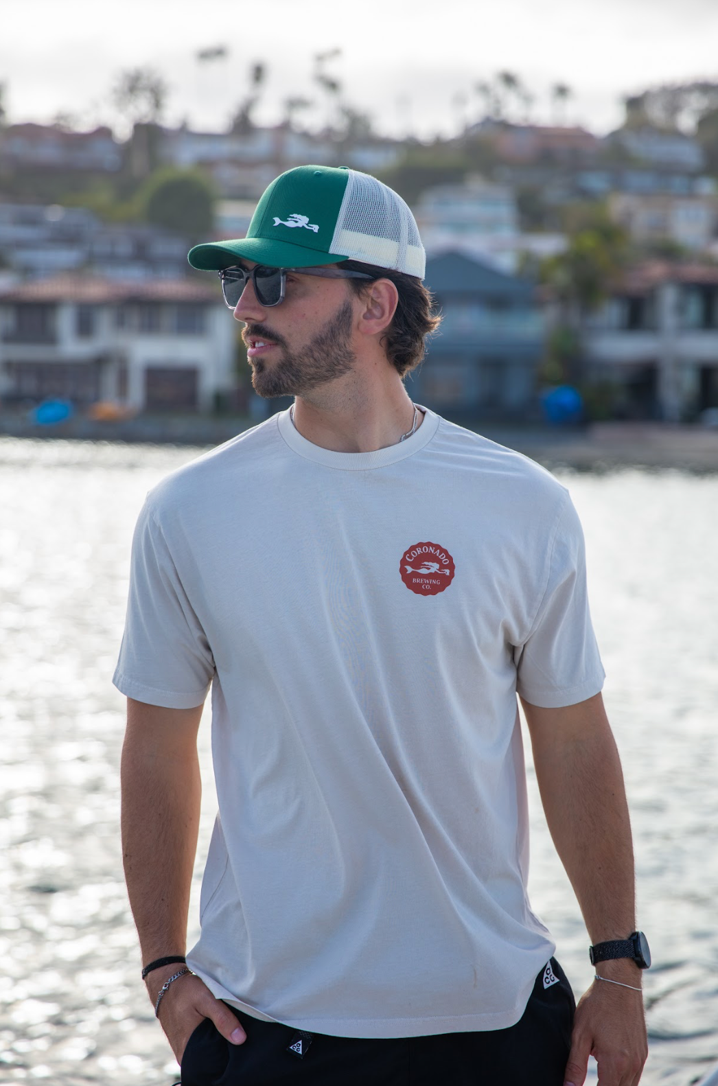 A man wearing a Coronado Brewing Co. t-shirt and a green and white cap, standing by a waterfront.