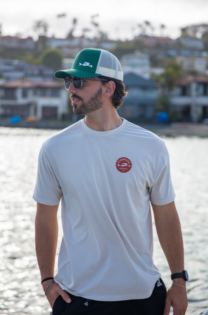 A man wearing a Coronado Brewing Co. t-shirt and a green and white cap, standing by a waterfront.