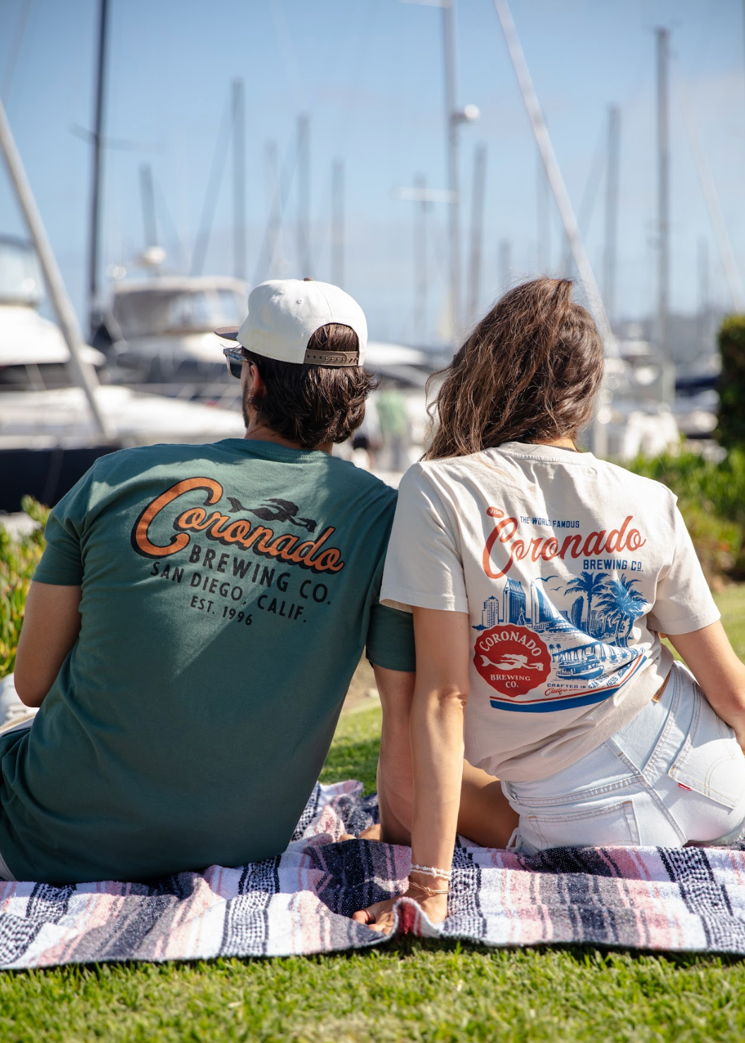 Two people sitting on a grassy area with marina in the background, wearing Coronado Brewing Co. t-shirts.