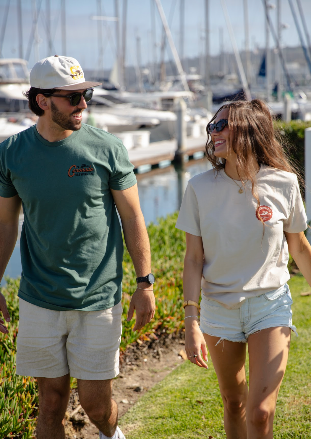 Two people walking wearing Coronado Brewing Co. apparel  by a marina with boats in the background